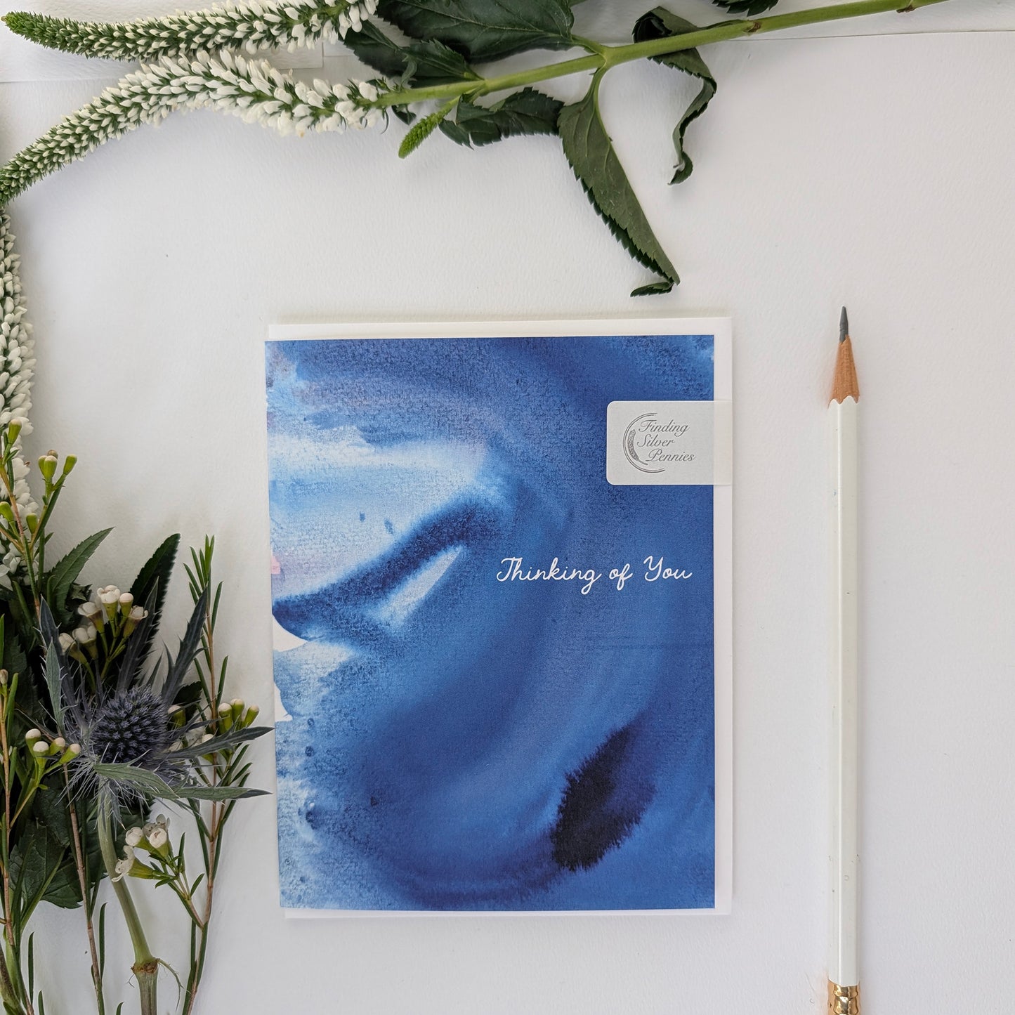Blue watercolor card with 'Thinking of You' text, surrounded by greenery and a pencil on a white surface.