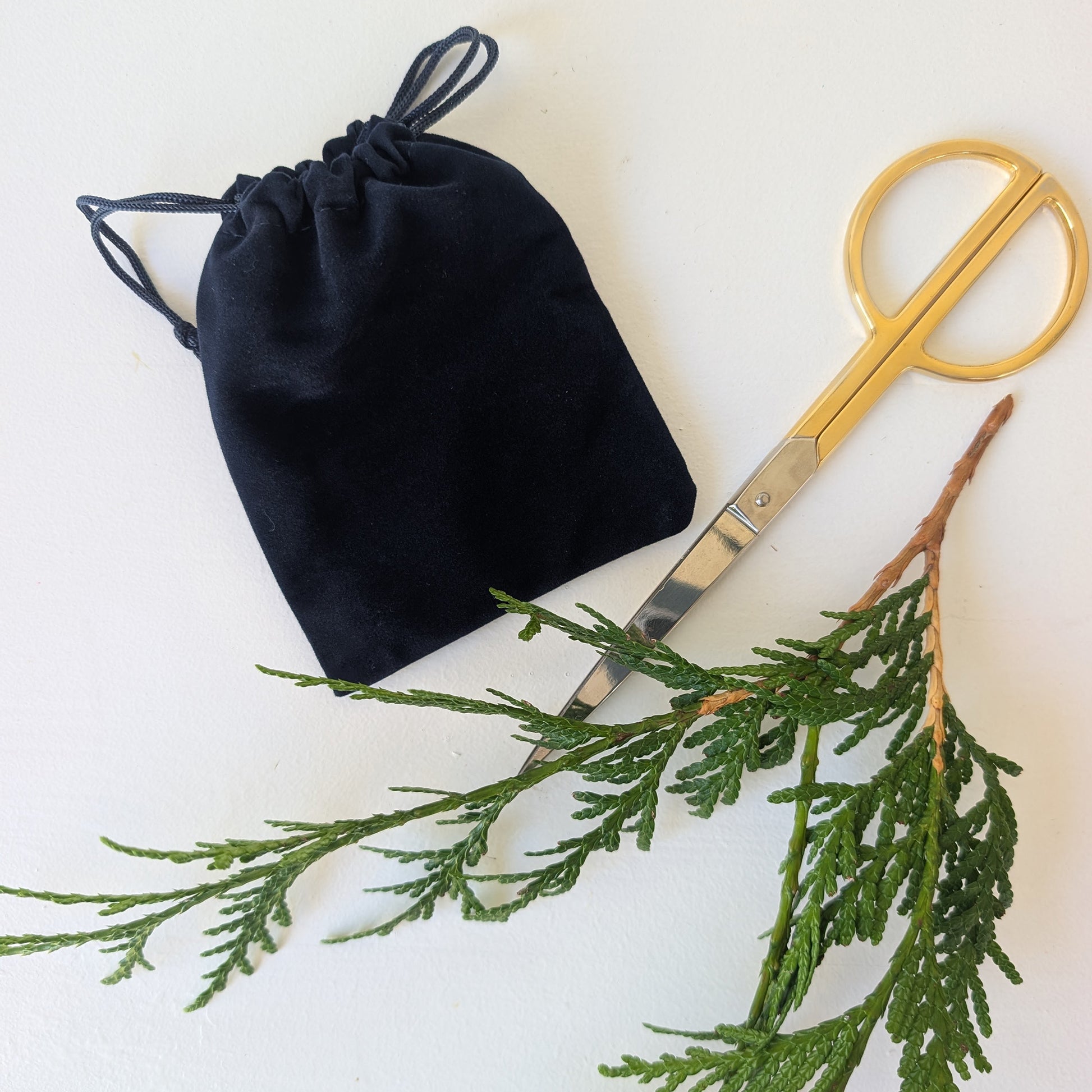 Navy blue drawstring velour bag, gold scissors, and green branches on a white background