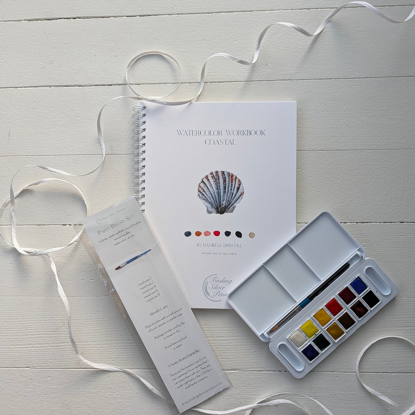 Watercolor workbook, color chart, and paint palette on a light wooden surface with ribbons.