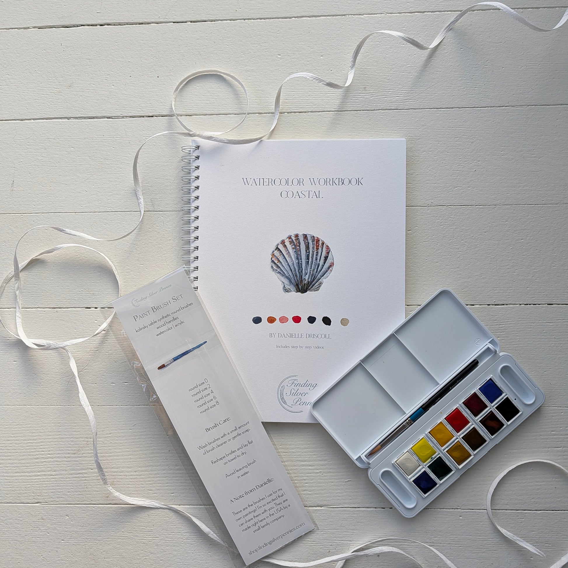 Watercolor workbook, color chart, and paint palette on a light wooden surface with ribbons.