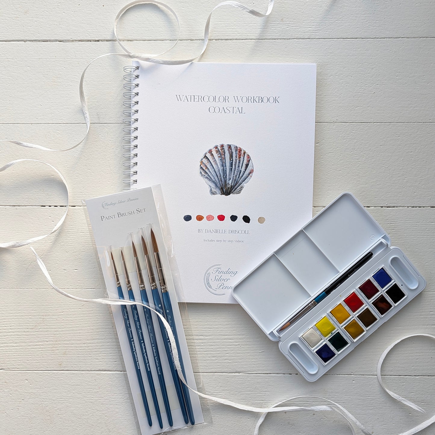 Watercolor painting set with a workbook, paintbrushes, and color palette on a light wooden surface.