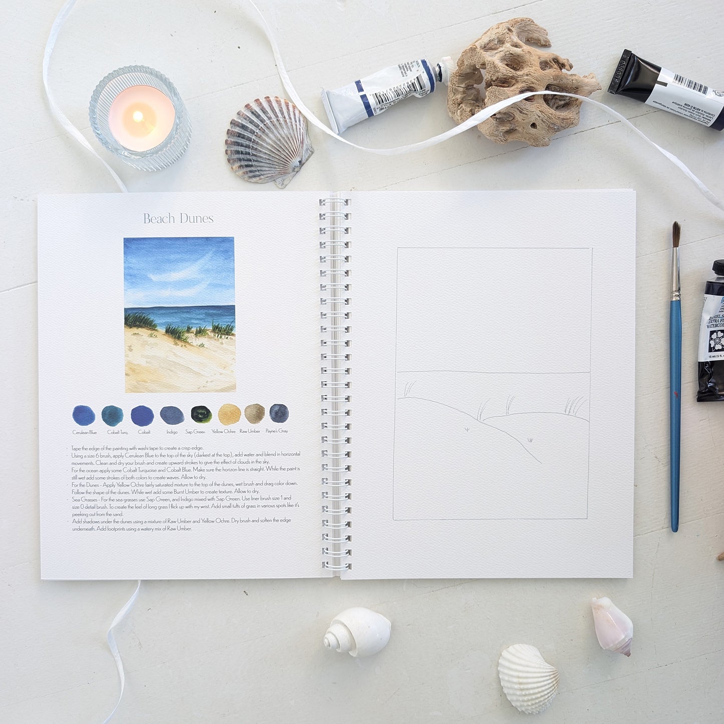 Open watercolor workbook with beach dune theme on page on a white surface with art supplies.
