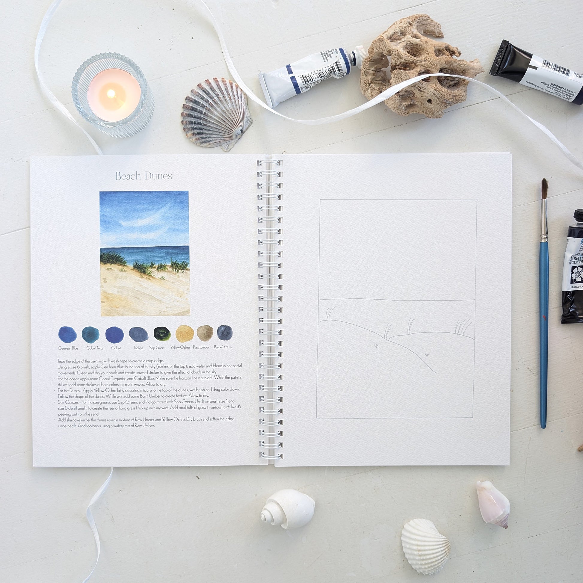 Open watercolor workbook with beach dune theme on page on a white surface with art supplies.
