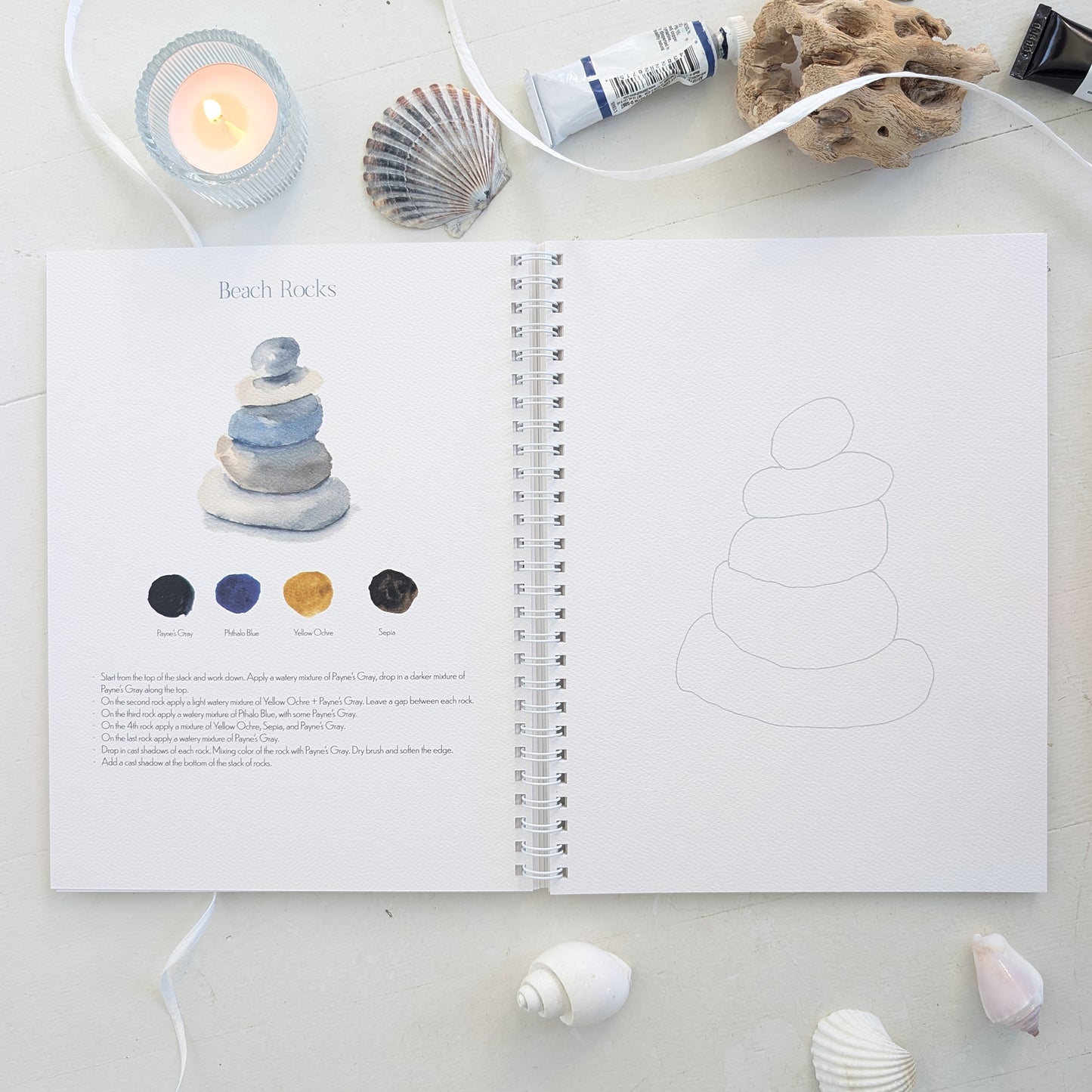 Open watercolor workbook with beach rocks on a light surface with decorative elements.