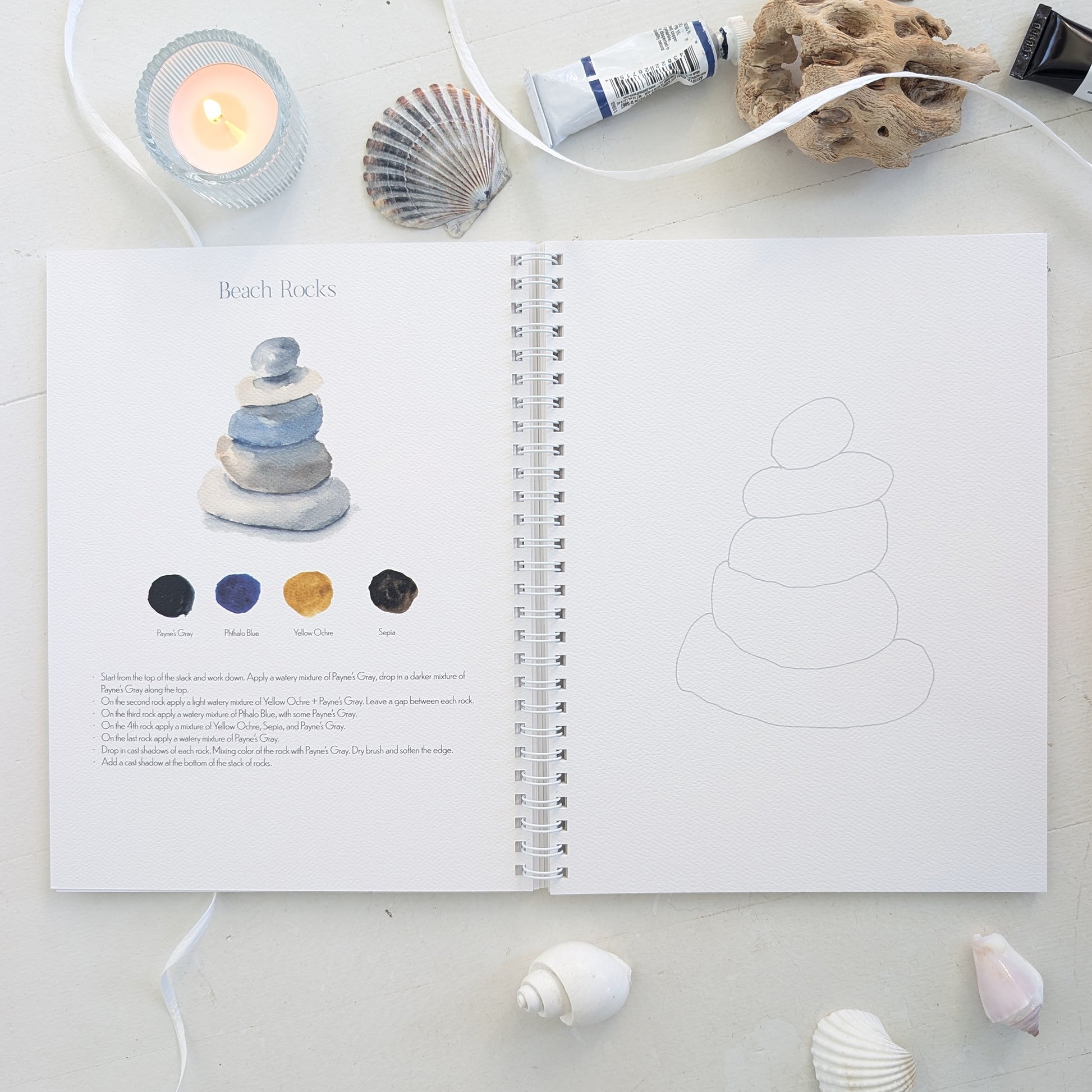 Open watercolor workbook with beach rocks on a light surface with decorative elements.
