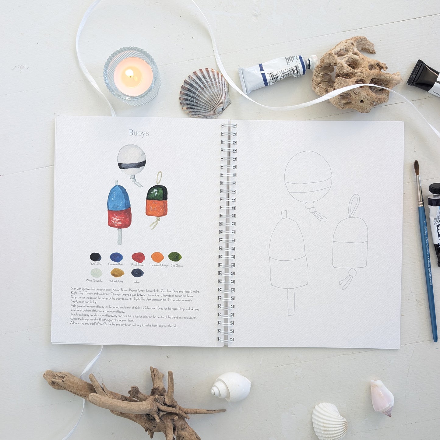 Open watercolor workbook with illustrations of buoys and color swatches on a white surface with decorative items.
