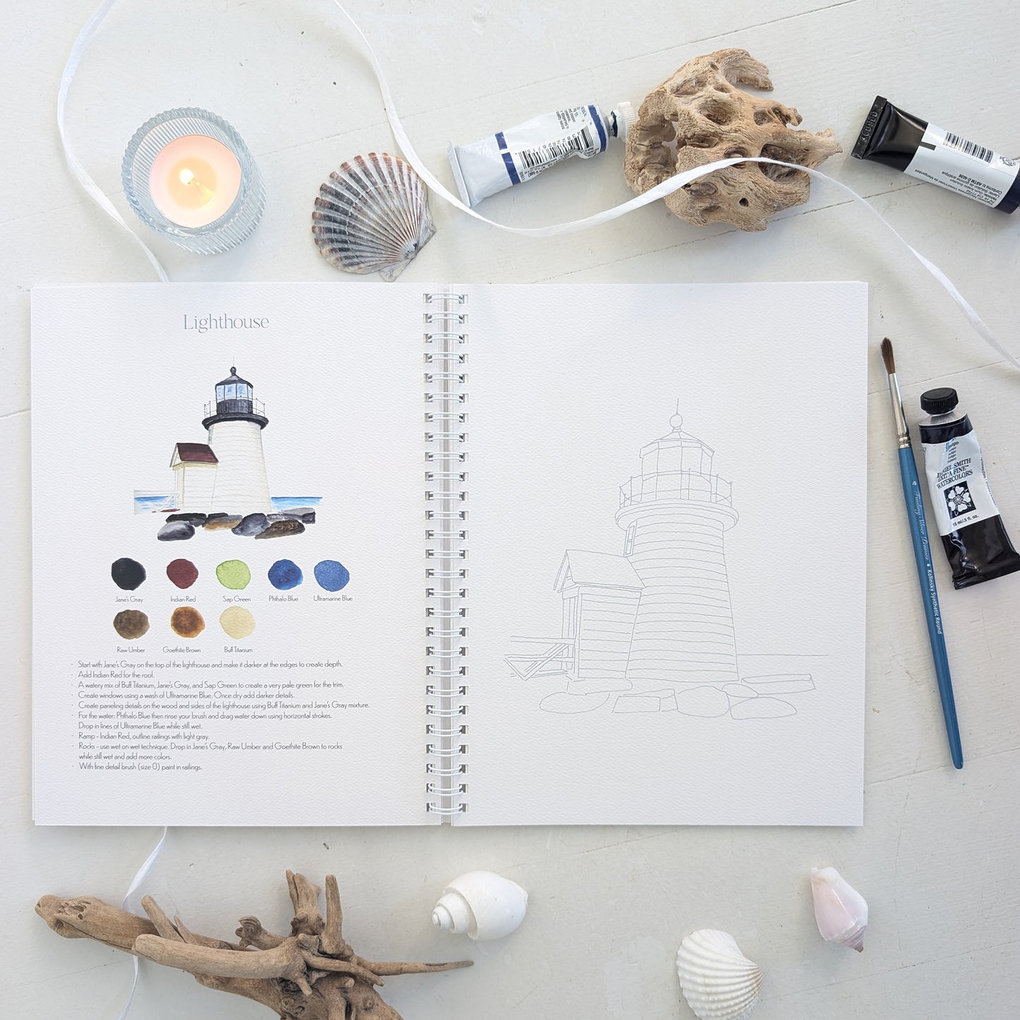Open watercolor workbook with a lighthouse illustration and color swatches, surrounded by art supplies on a light surface.