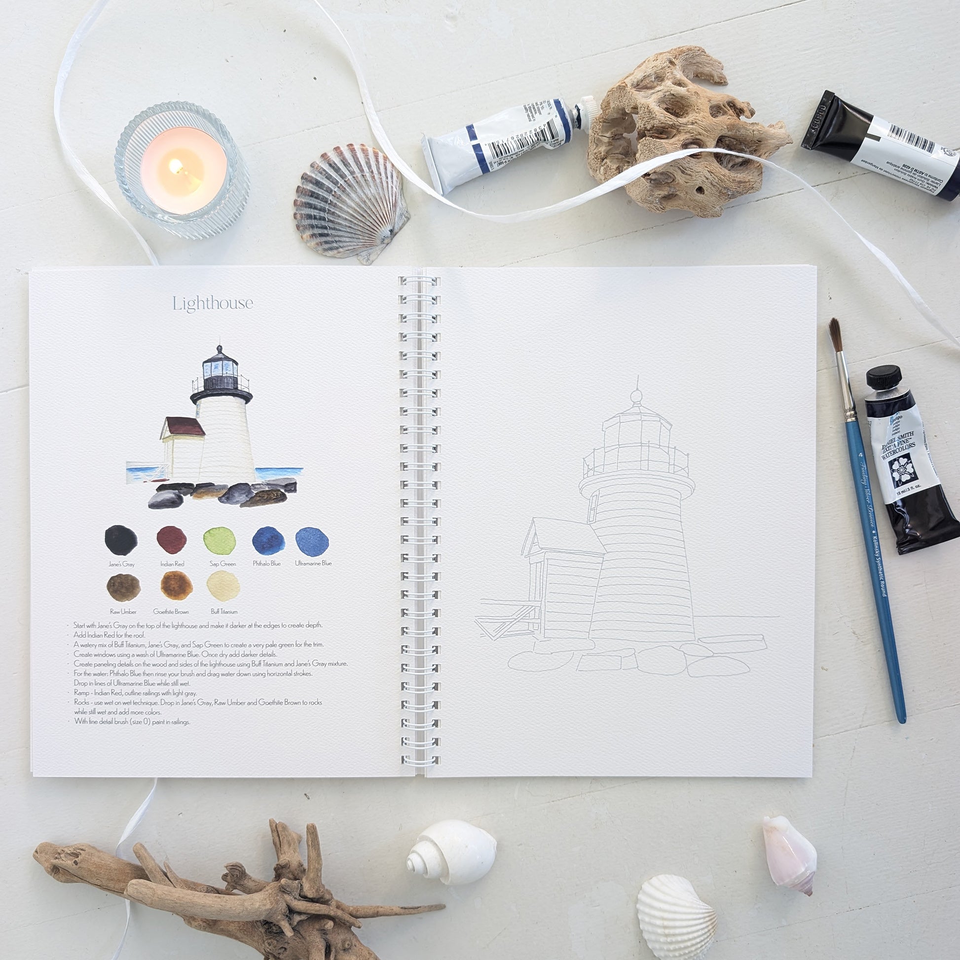 Open watercolor workbook with a lighthouse illustration and color swatches, surrounded by art supplies on a light surface.