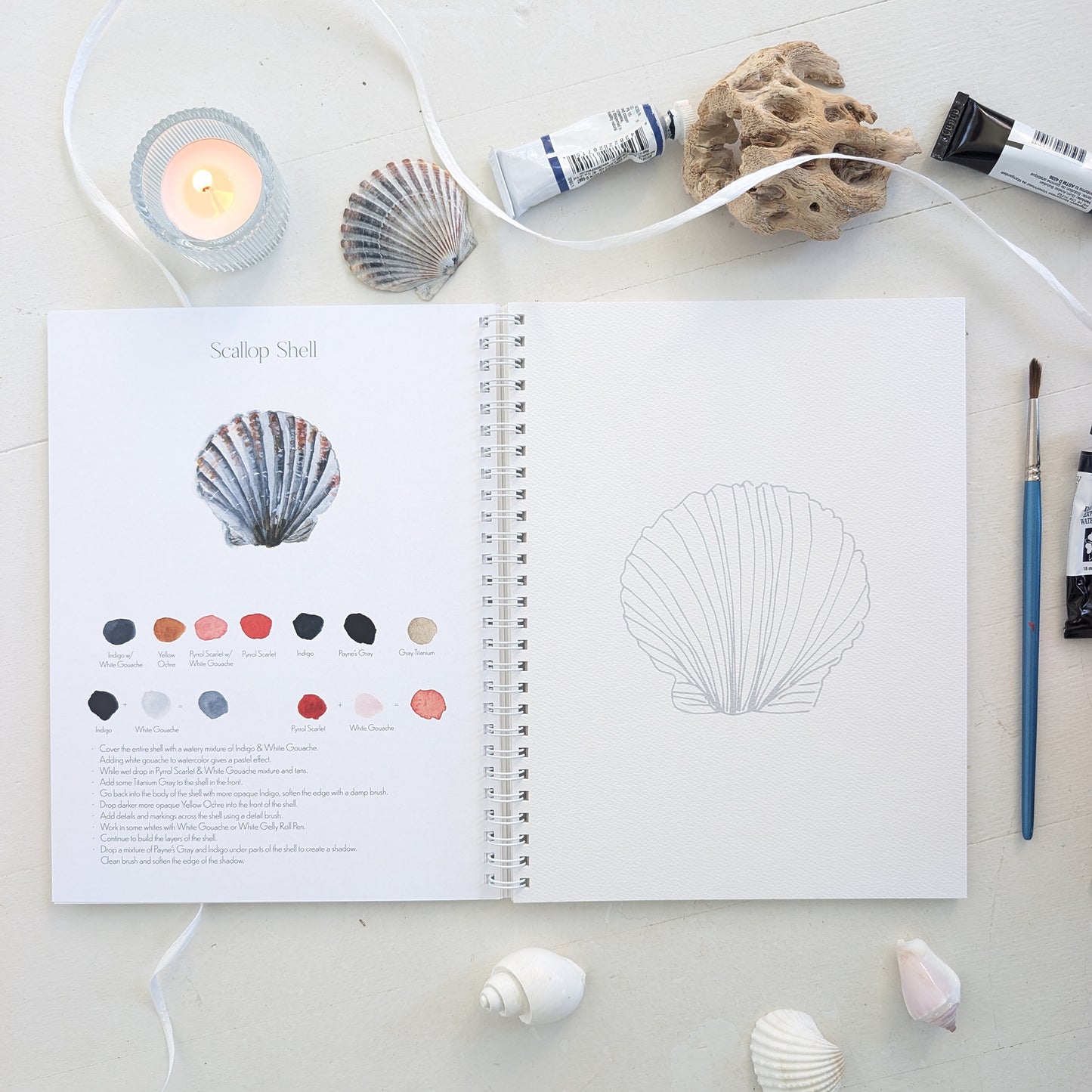 Open watercolor workbook with shell design and color swatches on a surface with seashells and a candle.