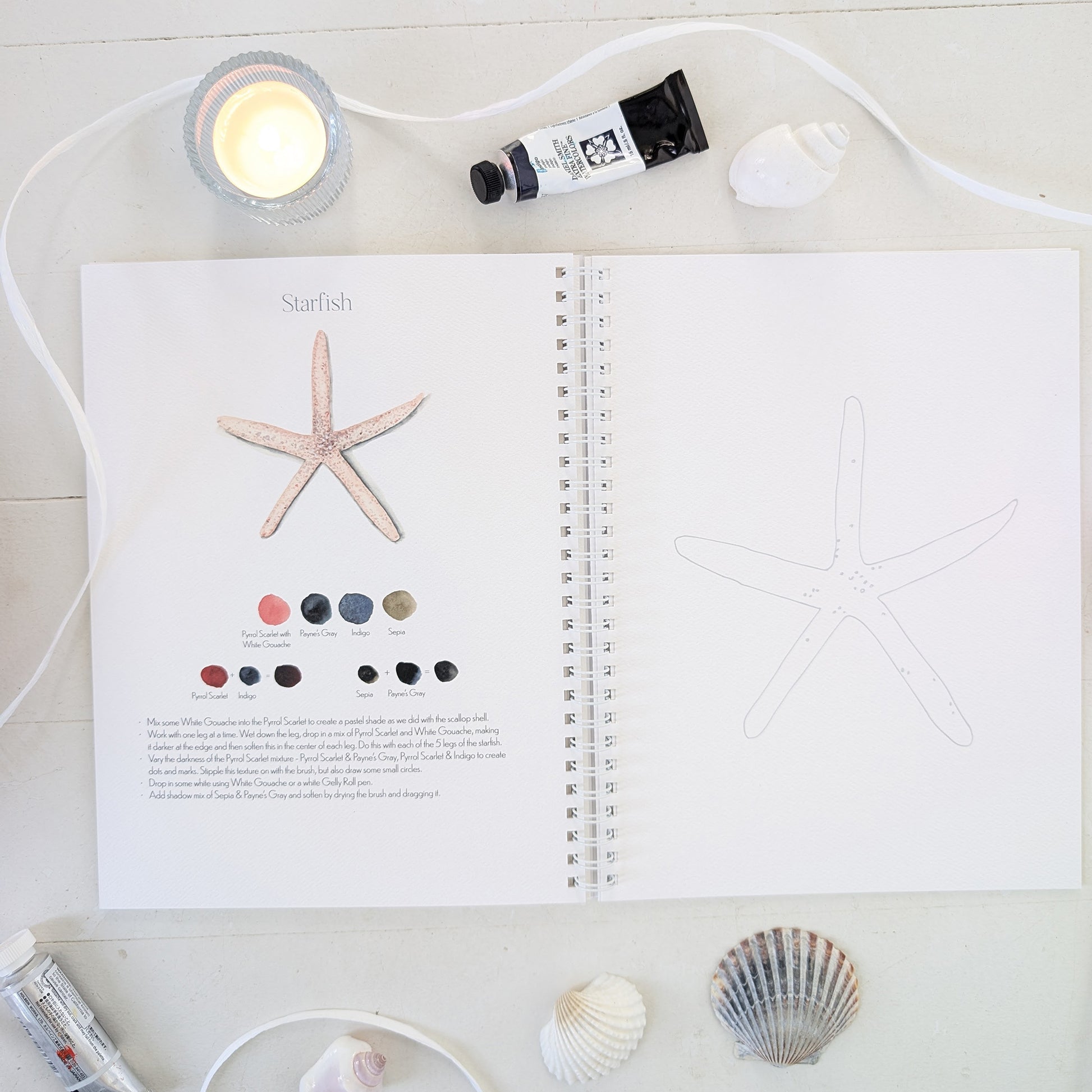 Open watercolor workbook with starfish illustration and color swatches on a white surface with a candle and shells.