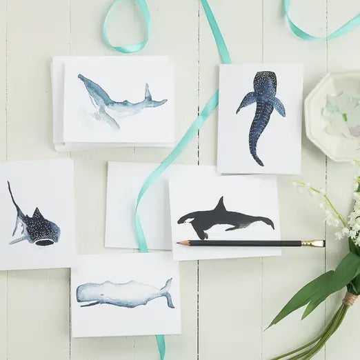 A styled flat-lay of white art cards featuring watercolor and silhouette whale illustrations, arranged on a white wood background with a mint ribbon, pencil, flowers, and a small dish.