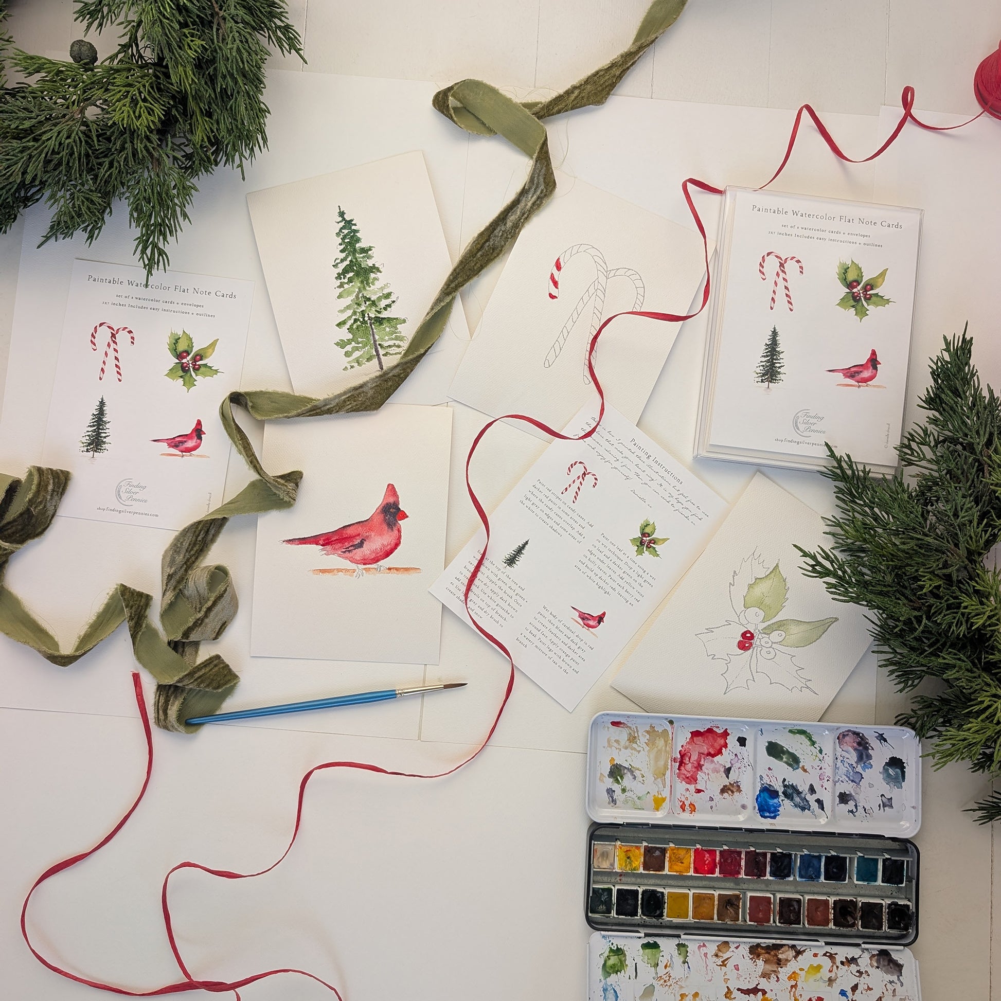 Holiday-themed cards with watercolor illustrations, ribbons, and a paint set on a white surface.