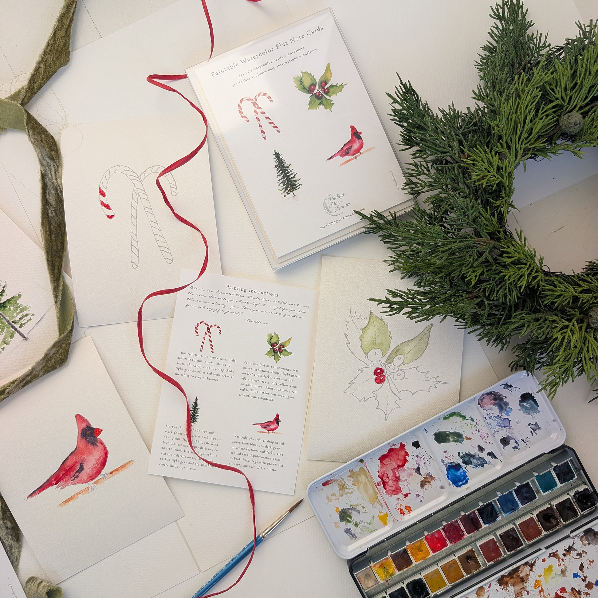 Art supplies including watercolor paints, brushes, and Christmas-themed cards on a white surface.