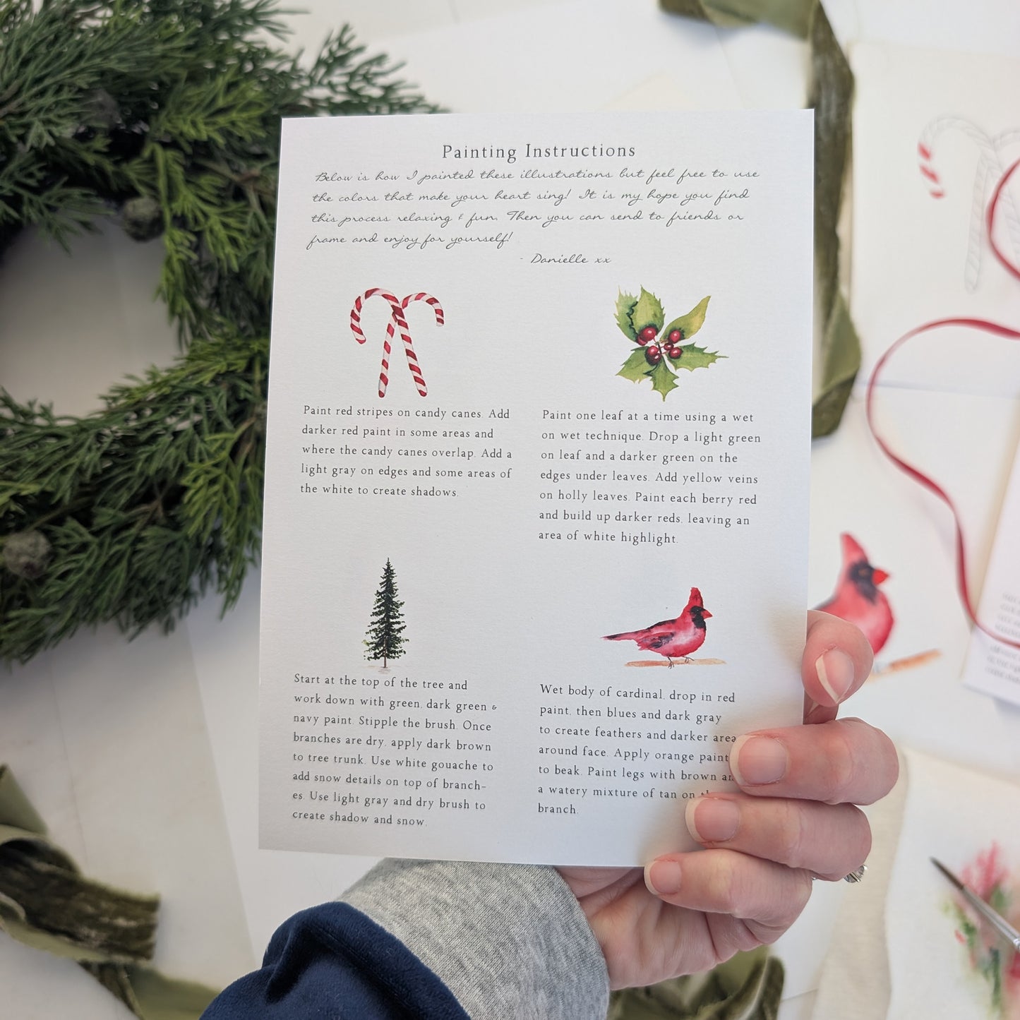 Hand holding a card of painting instructions with Christmas-themed illustrations on a white surface with greenery.
