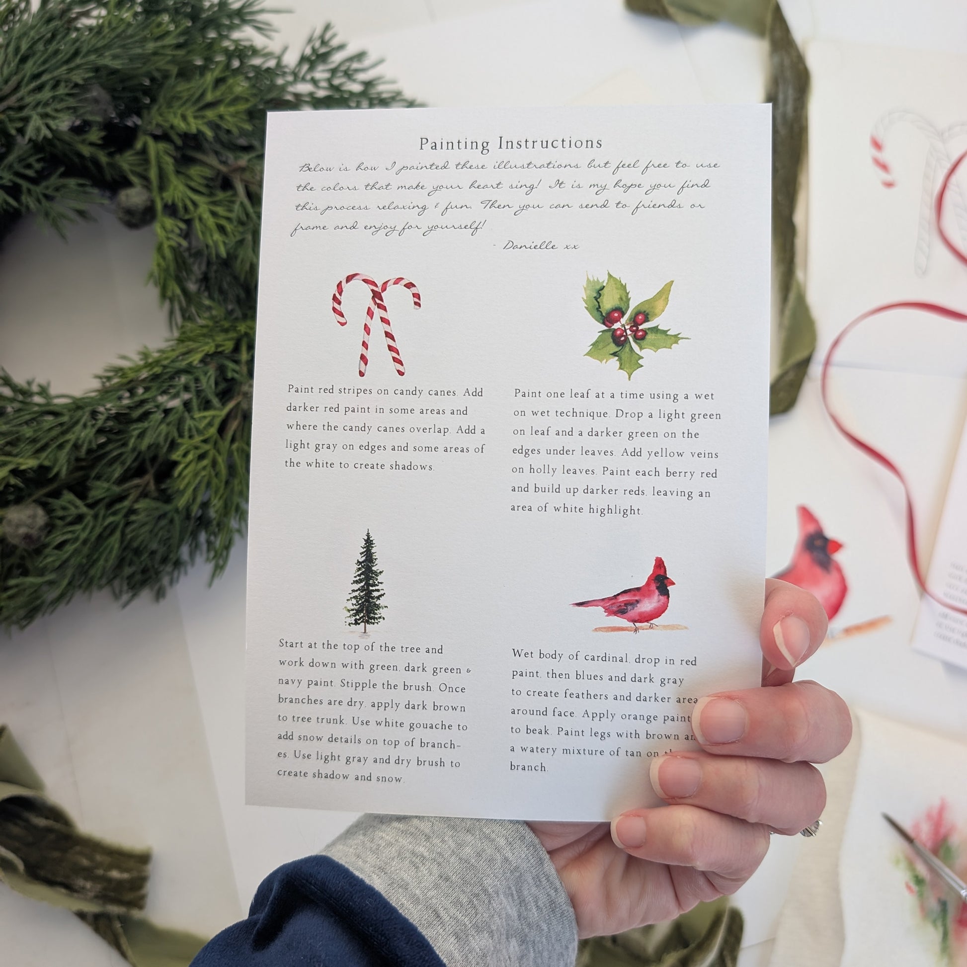 Hand holding a card of painting instructions with Christmas-themed illustrations on a white surface with greenery.