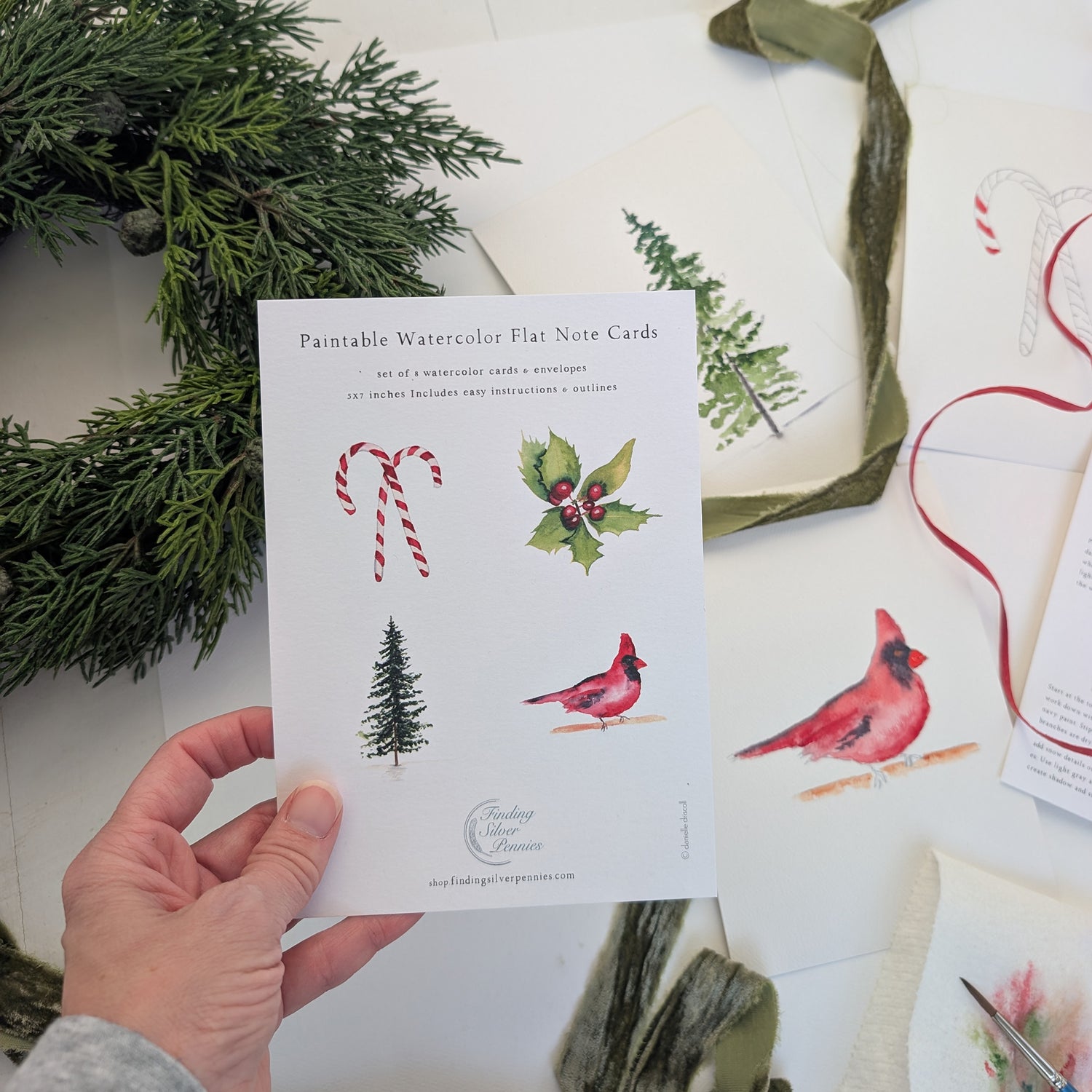 Hand holding a pack of paintable watercolor flat note cards with Christmas designs on a white surface with greenery.