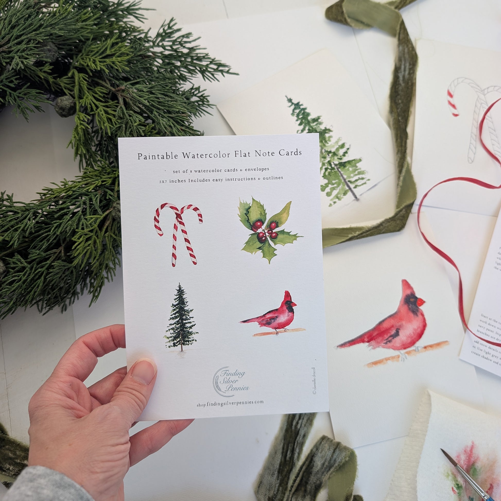 Hand holding a pack of paintable watercolor flat note cards with Christmas designs on a white surface with greenery.