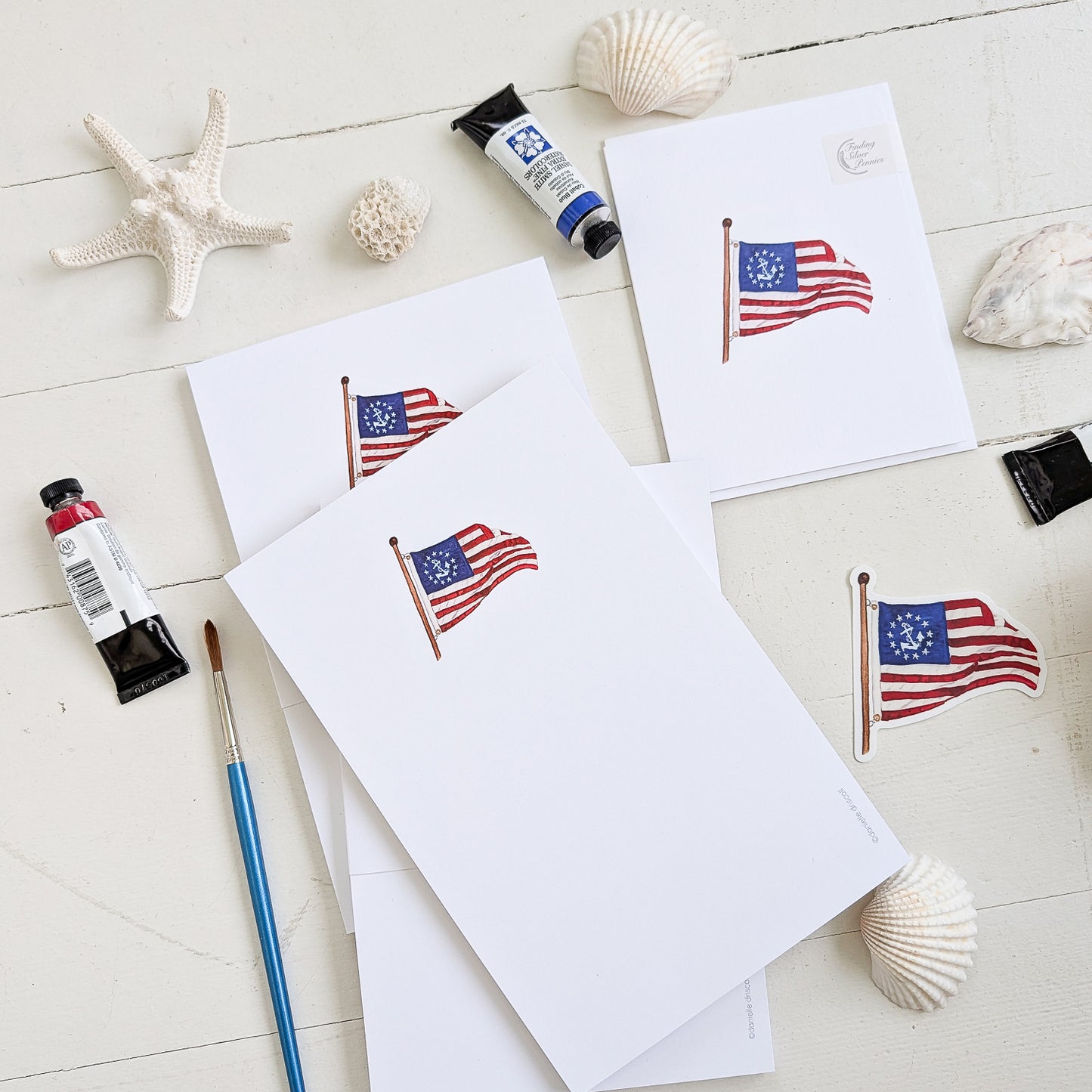 Notepads and cards with American Yacht Ensign flag design on a light wooden surface with paint tubes and brushes.