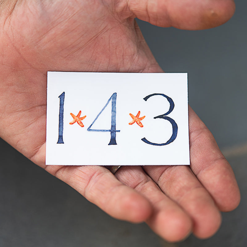 Hand holding a 143 sticker. Original painting by Danielle Driscoll of Finding Silver Pennies
